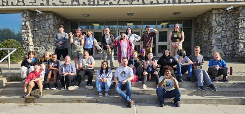 A group of people sit and stand outside a building, posing and smiling for a group photo.